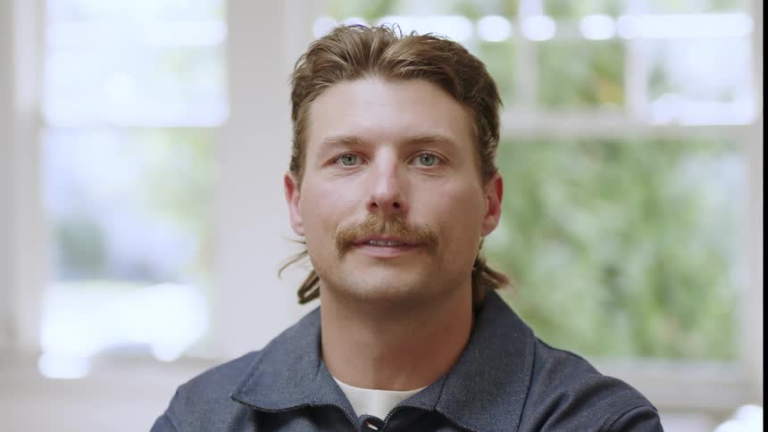 Close up of the face of a man with long hair and a mustache looking at the camera