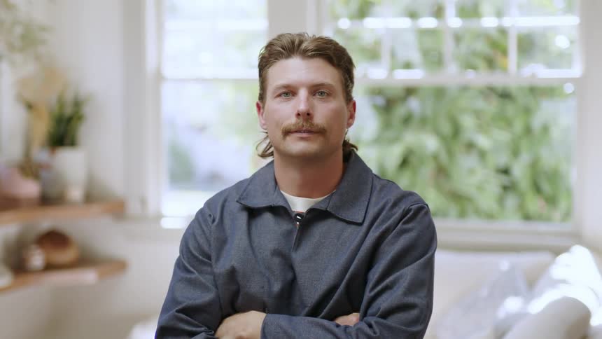 A man with a mustache and long hair sitting on the living room looking at the camera
