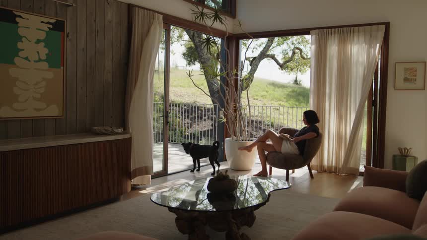 A young woman sits on a chair with a dog nearby looking at the backyard through large windows