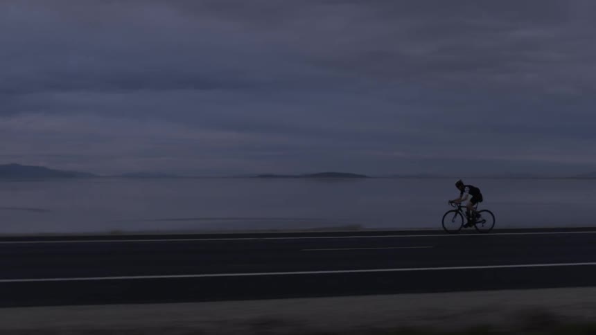 Person riding bicycle on highway next to lake on cloudy day after sunset