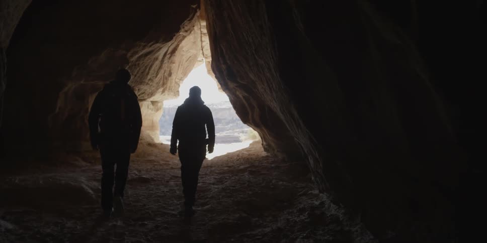 Two people walk through a rock crevice toward the sunlight