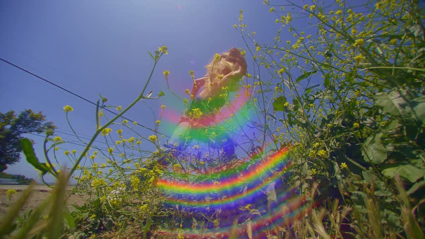 A woman dances on a patch of wildflowers under a clear blue sky on a sunny day with iridescent colorful reflections