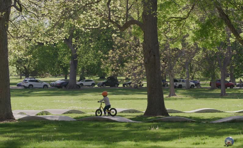 Little boy riding a bicycle with a helmet in a park surrounded by trees and cars parked in the background
