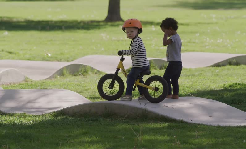 A child wearing a helmet rides a yellow bike on a wavy path in a park with another child standing to the side
