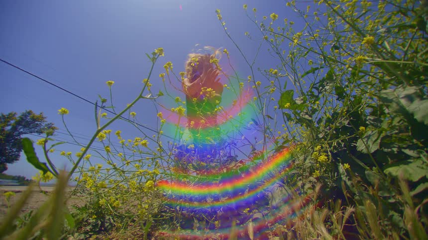 A woman dances on a patch of wildflowers under a clear blue sky on a sunny day with iridescent colorful reflections