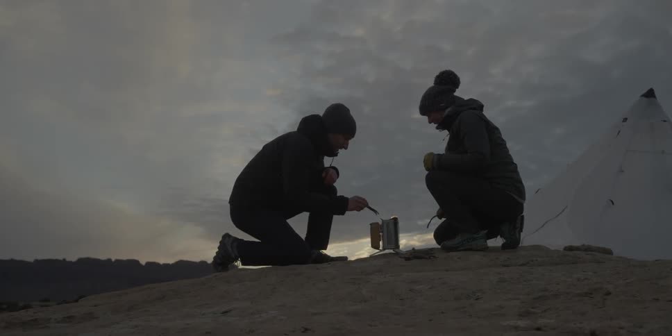 Two people huddle around a small fire in a canister at dusk