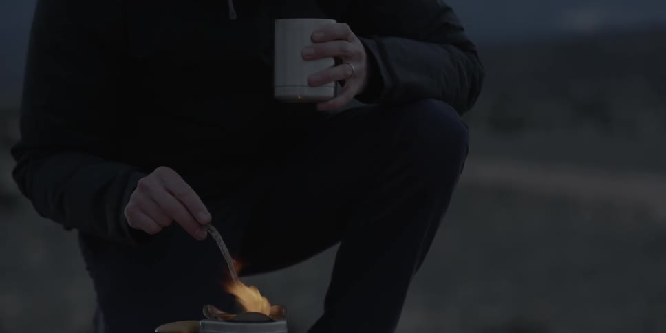 A man is cooking in a small pot holding a mug in the wilderness