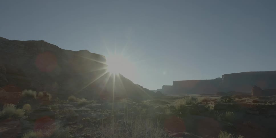 The sun rises and peeks out around the side of a cliff onto the desert