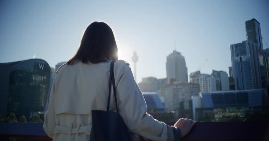 A woman has her back turned and is observing the buildings of a city and strong sunlight coming from the sky