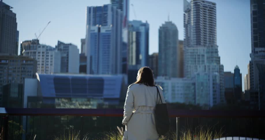 A woman walks towards a railing looking at a city skyline with various buildings and construction cranes
