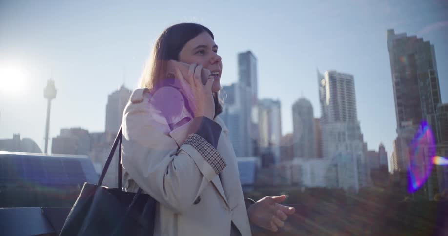 A woman wearing a coat outdoors in a city places a phone call using a smartphone