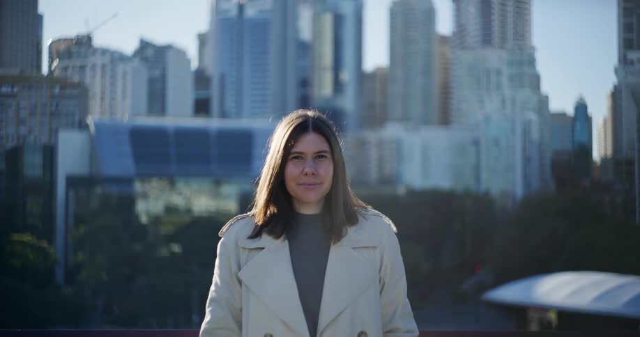 A woman in a white coat is standing staring straight ahead and behind her at huge buildings in a metropolitan city