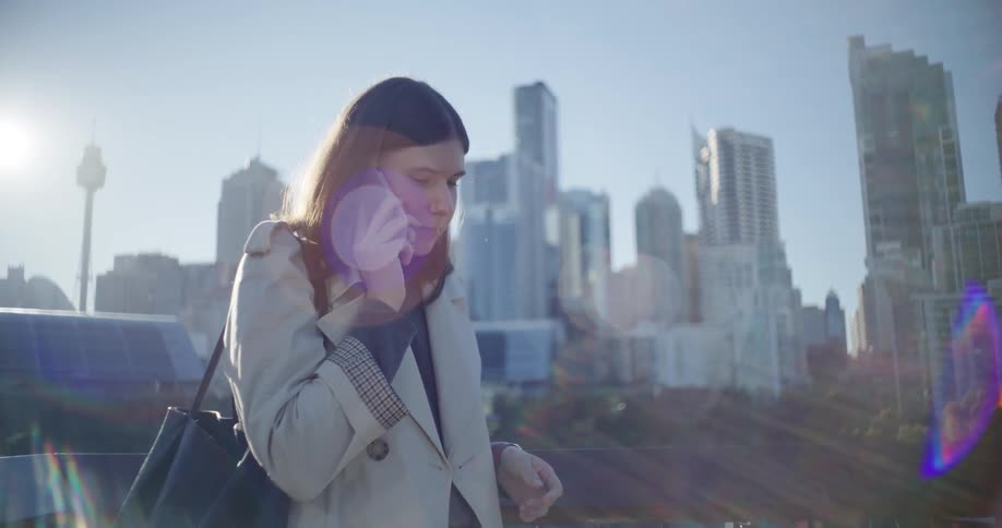 A woman holds a smartphone answering a call with a city skyline in the background on a sunny day