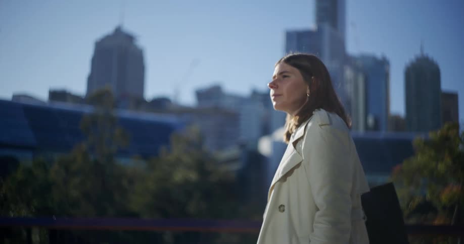 A woman with long hair wearing a coat walks gazing at a city skyline with skyscrapers