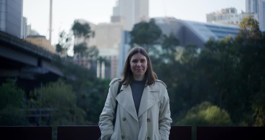 A white woman wearing a coat and carrying a bag smiles in front of a downtown city area