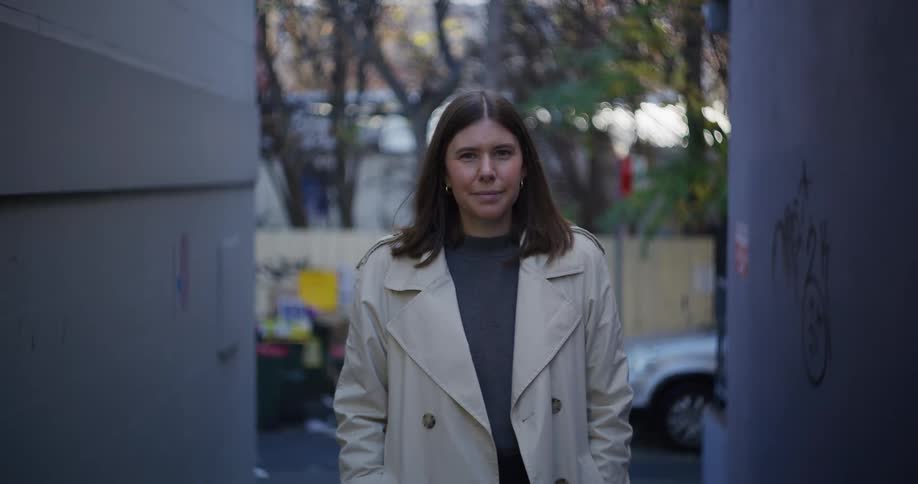 Woman in a white coat giving a slight smile while standing in an alley in a city
