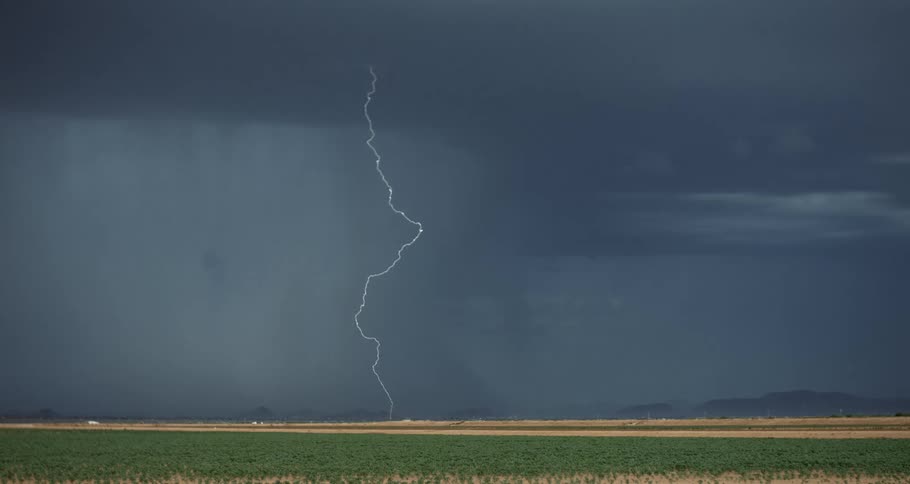 Lightning bolts zig zagging through a gray sky during a thunderstorm over farm land