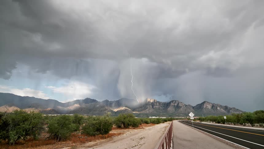 Storm clouds loom over a road leading towards mountains as lightning flashes in the sky