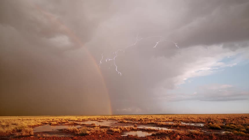 A storm dropping lightning by a rainbow over a grassy area