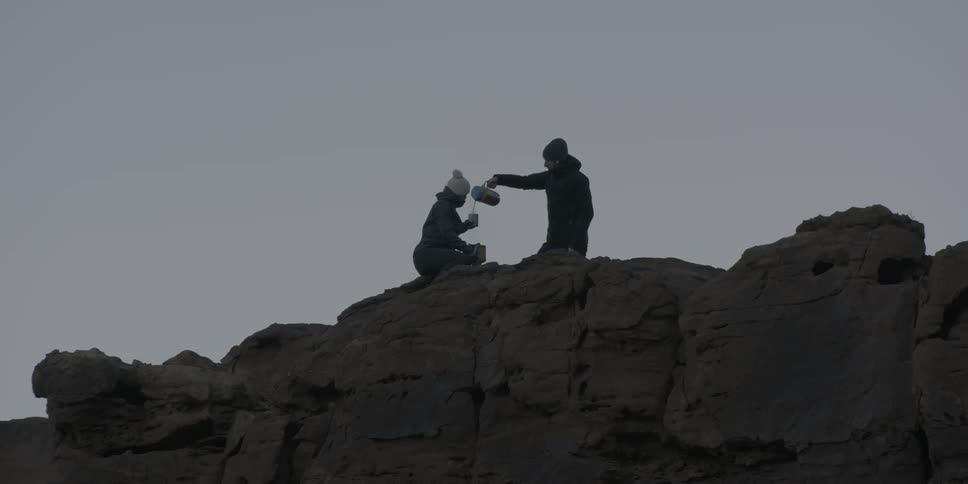 While sitting at the top of a cliff, one person pours a liquid into the cup of the other person