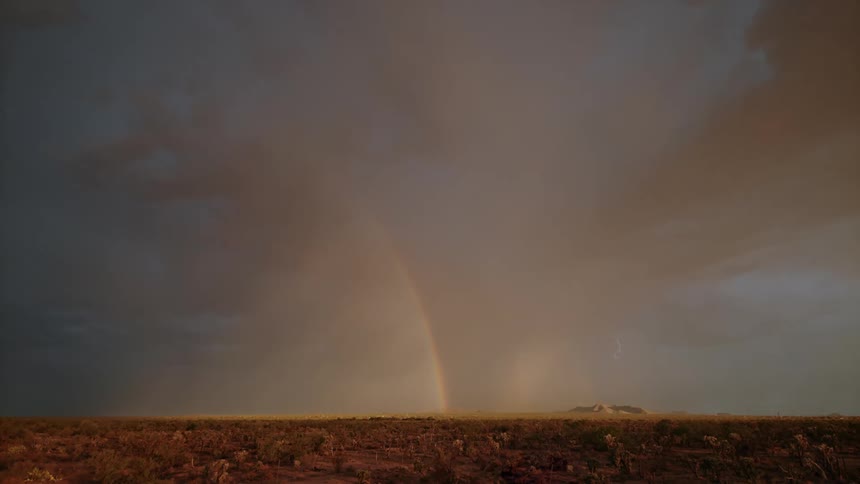 Dark storm clouds emit lightning near a rainbow arcing down to a grassy area