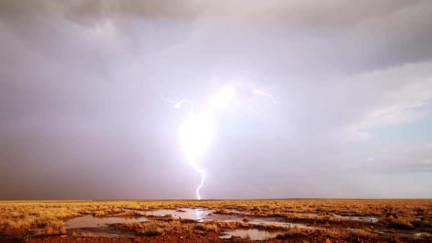 A lightning bolt strikes the ground next to a rainbow over a vast landscape during a storm at daytime