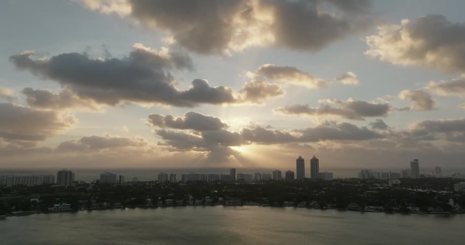 A coastal city skyline during sunrise or sunset with the sun partially obscured by clouds