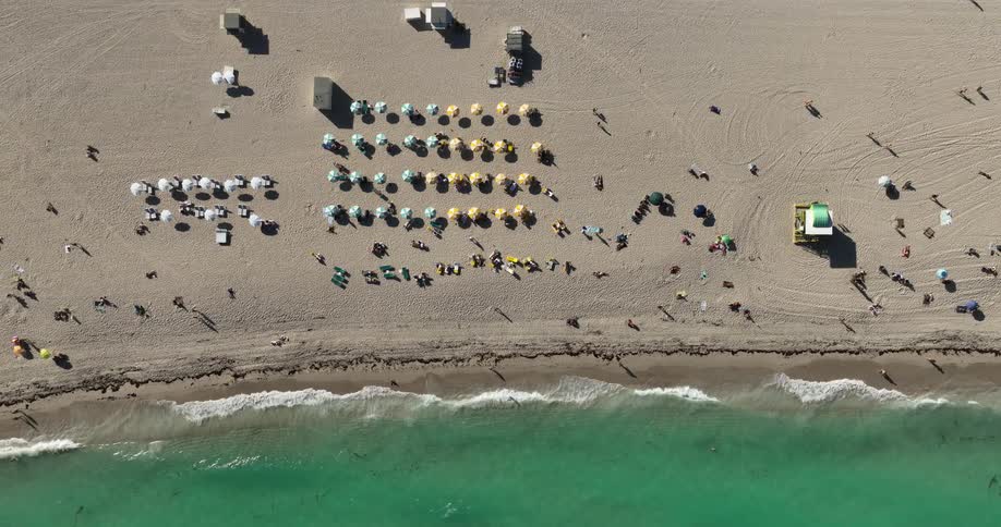An aerial view of a sandy beach with scattered people some sitting under parasols arranged in rows