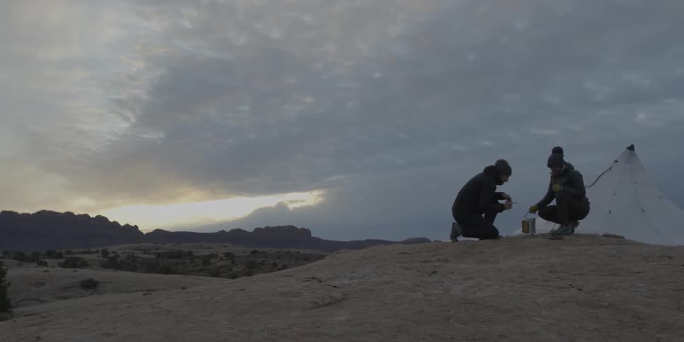 Two people crouch near a small canister sitting on the ground at dusk