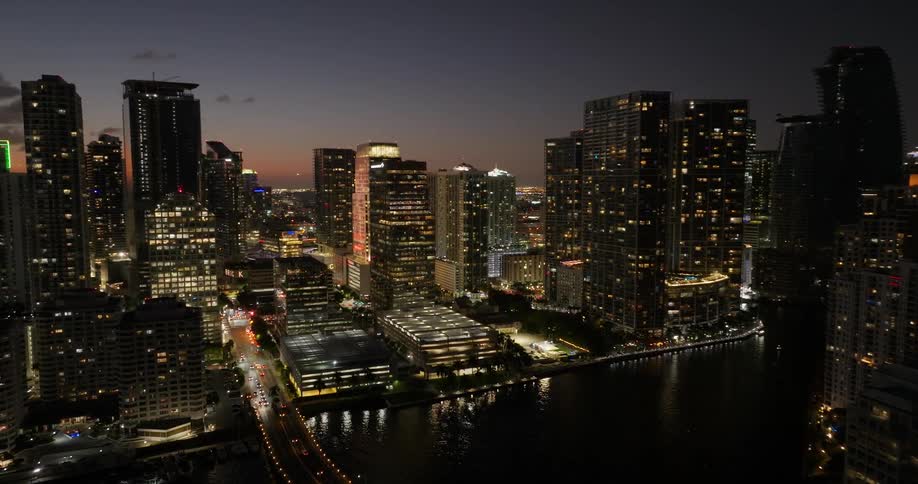 An aerial view of a metropolitan city at night with the building lights on and there is a large bridge