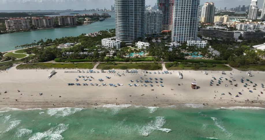 An aerial view of a beachfront with waves approaching the shore blue beach umbrellas and sun loungers neatly arranged in front of the city