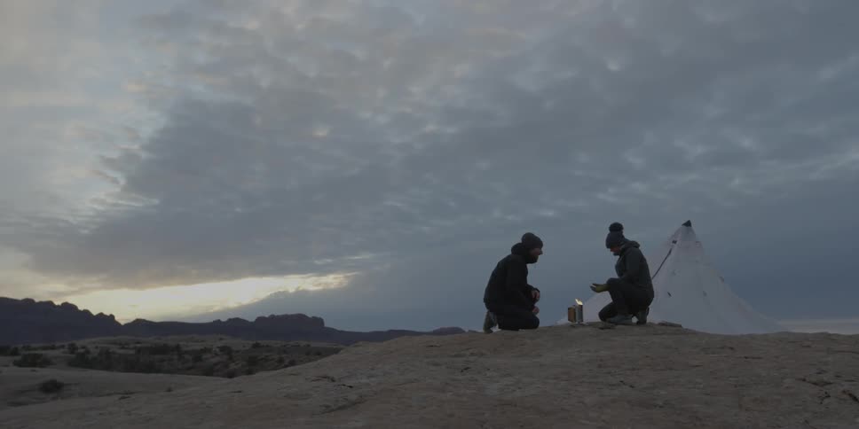 Two people are camping on a small hill at sunset