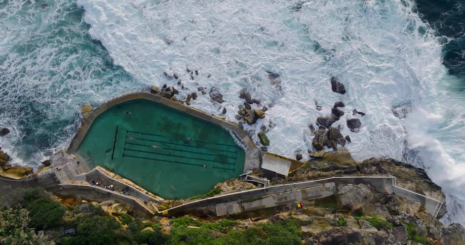 People swim in a swimming pool overlooking the ocean while waves crash against coastal boulders