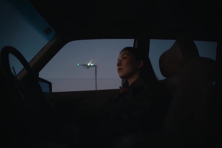 Woman sits in the passenger seat of a parked car at night as an airplane passes overhead