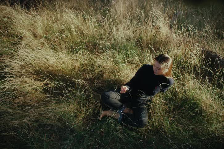 A young woman sits in tall grass playing with a flower