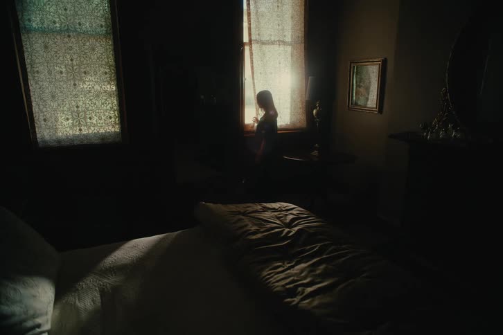 A person is in a dark room opening the curtain of a window where there is sunlight coming in