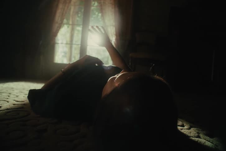 A woman lying on top of a bed her hand covering the sunlight coming in from the window