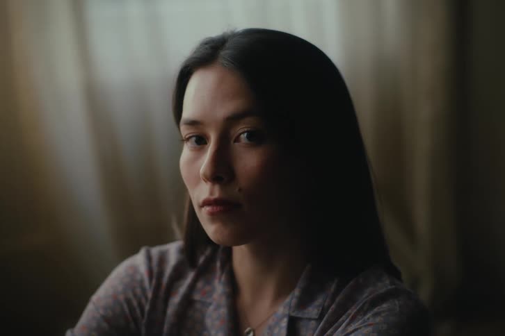 Woman near a curtained window looks around briefly before staring