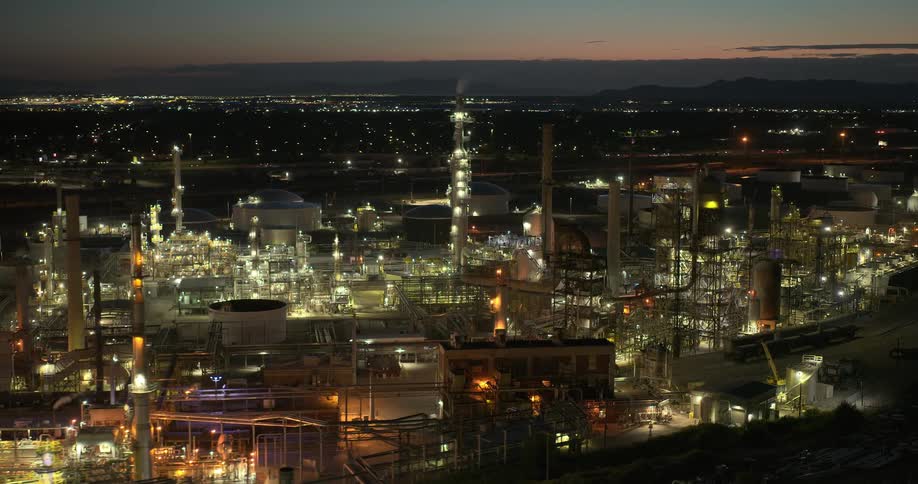 A large refinery lit up at dusk sits on the edge of a large city