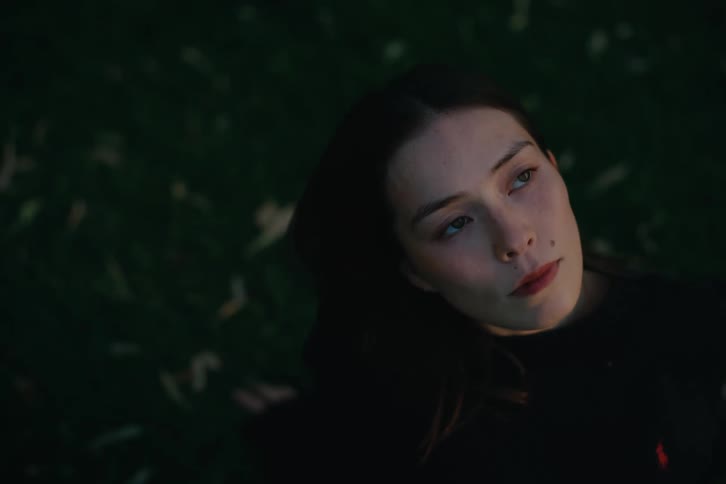 A dark haired woman sitting on grass looks contemplative