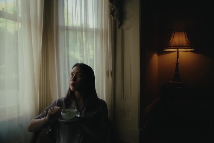 A woman sitting by a window with sheer curtains holding a cup with a lamp on to the side casting a warm glow