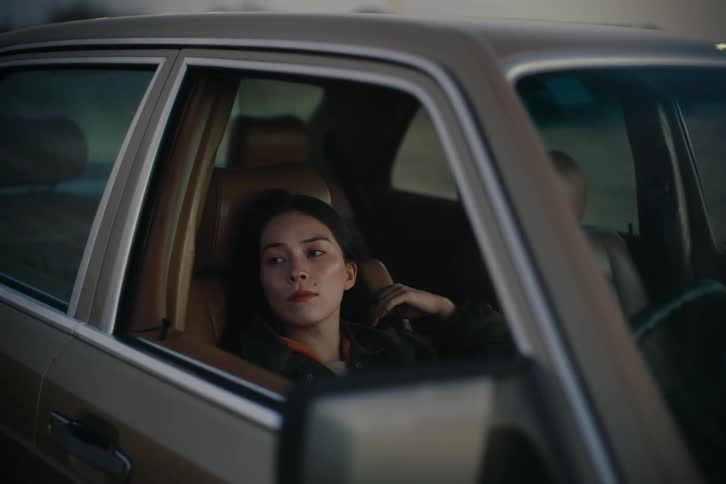 A dark haired woman brushes her hair with a hand while sitting in the passenger seat of a car