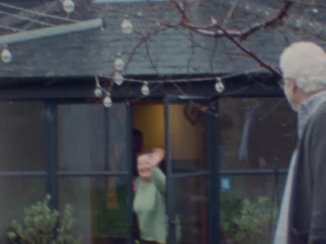 An elderly man is in a back yard as an elderly woman comes out of the house and calls for him waving as they both laugh