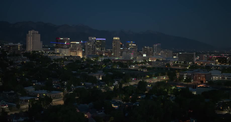 A nighttime cityscape with illuminated buildings against a mountainous backdrop