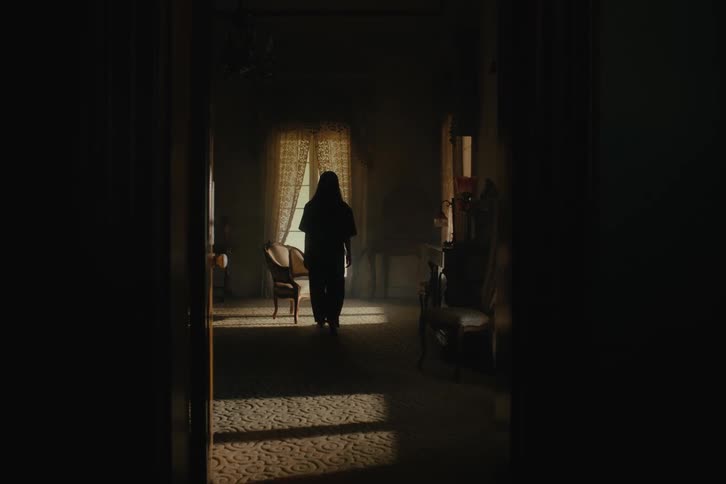 A person has their back turned and walks to a chair in a room that is a little dark and sits down