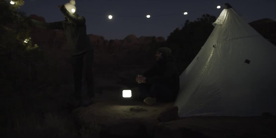 Woman hanging lights from tree to a tent while man sits and watches