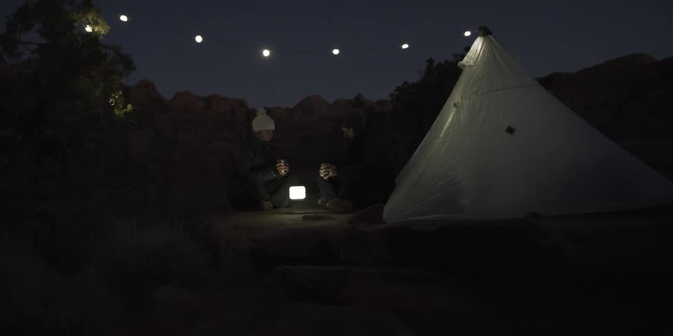 A couple sits in front of their tent under a string of lights