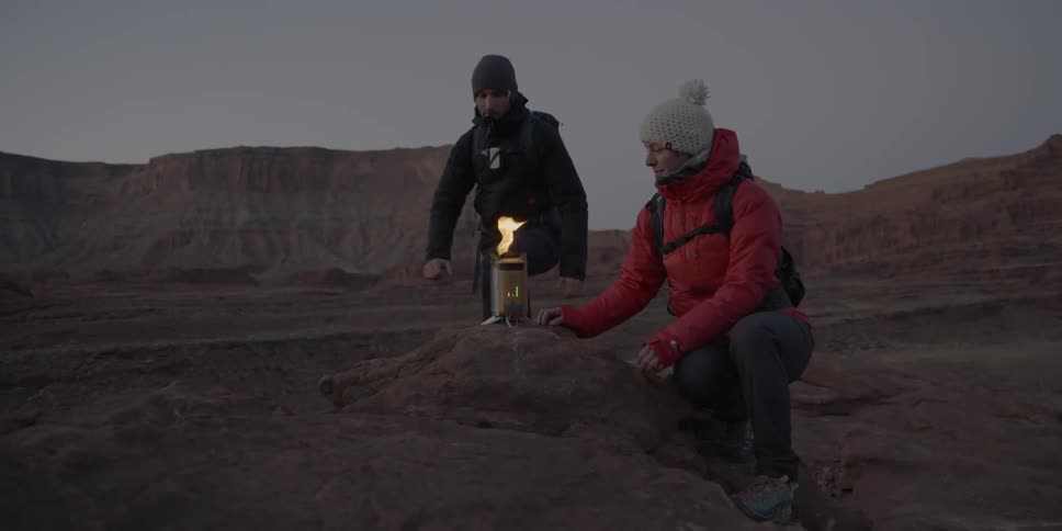 Two people have a small fire burning in a canister in the desert