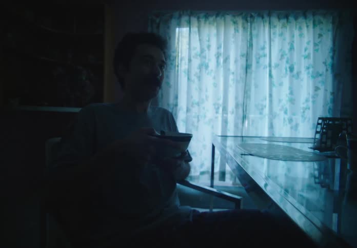 A man eating cereal in a bowl talking to a woman who is in front of him and they are both sitting in a kitchen