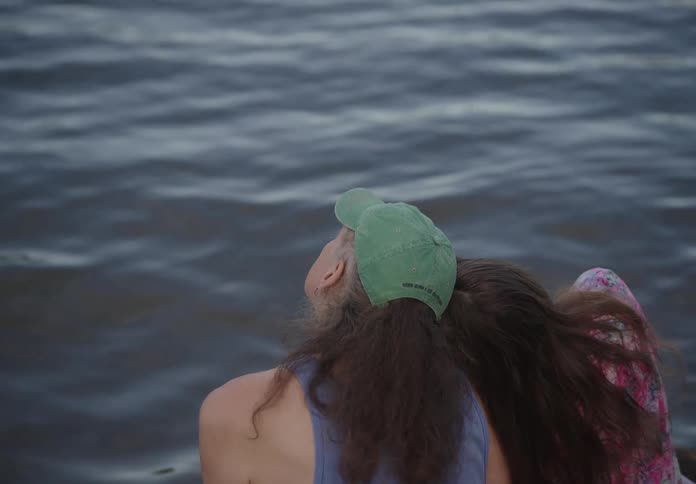 A person wearing a green cap leans their head on a person wearing blue shirt as they gaze out over a body of water enjoying the view